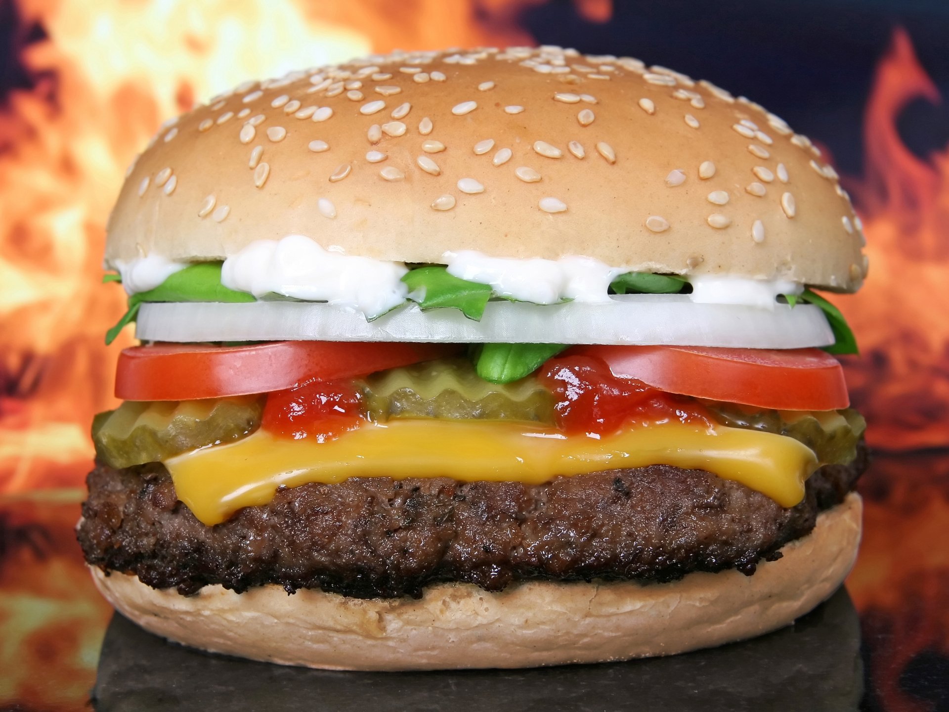 HD PC desktop wallpaper featuring a close-up of a juicy cheeseburger with lettuce, tomato, pickles, onions, ketchup, and mayonnaise against a fiery background.