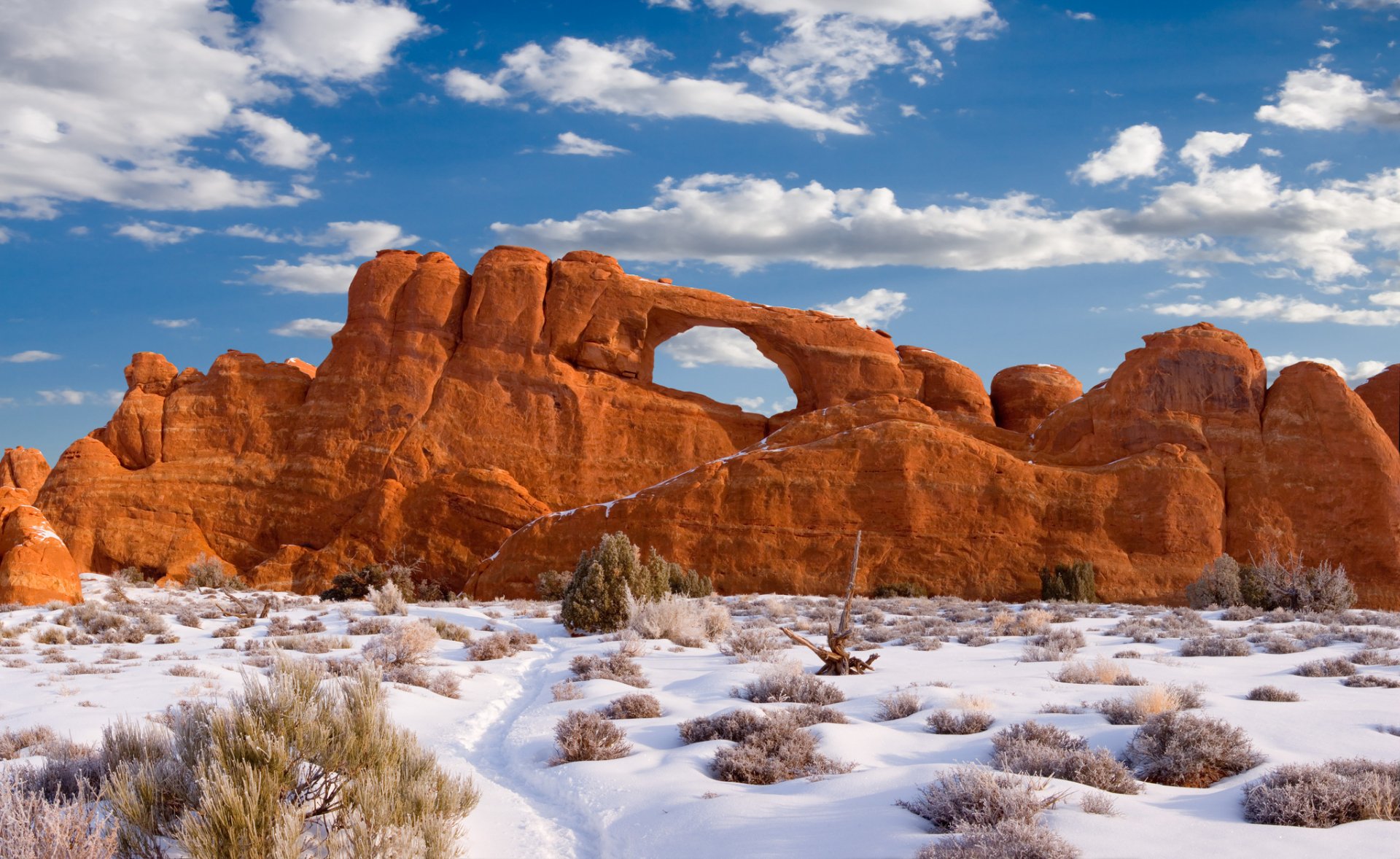 Stunning red rock formations rise against a blue sky, with wispy clouds above. A snowy landscape surrounds the iconic arch, showcasing the beauty of nature and landscape.