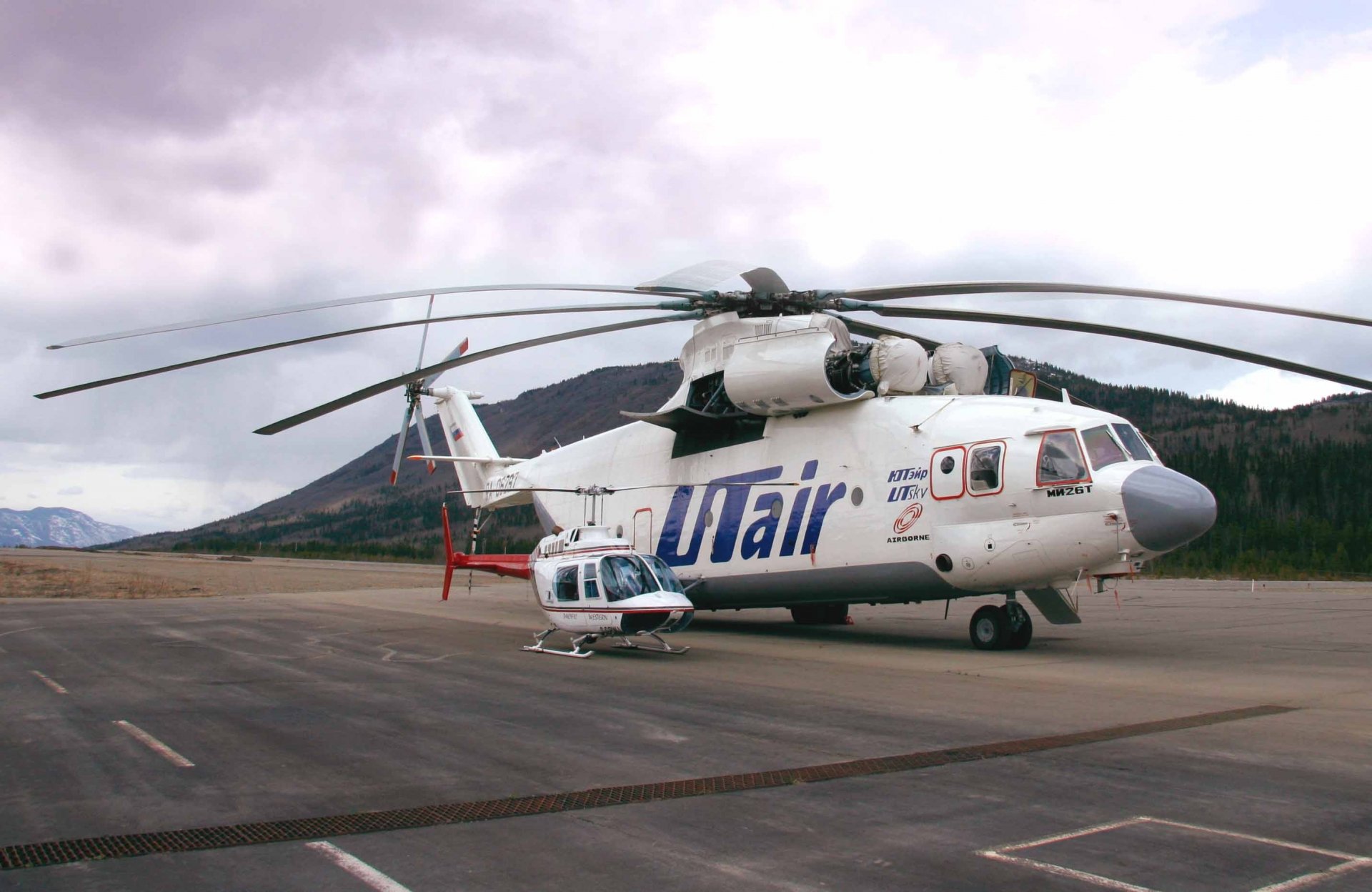 HD desktop wallpaper featuring a Mil Mi-26 helicopter with Utair livery on a tarmac surrounded by mountainous terrain under a cloudy sky.