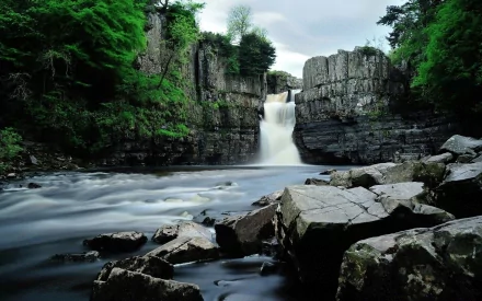 A serene nature scene featuring a waterfall cascading over stone cliffs, surrounded by lush green trees, flowing river, and smooth rocks, captured in a stunning time-lapse effect.