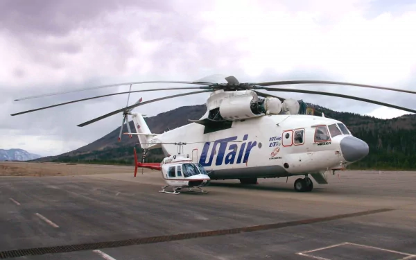 HD desktop wallpaper featuring a Mil Mi-26 helicopter with Utair livery on a tarmac surrounded by mountainous terrain under a cloudy sky.