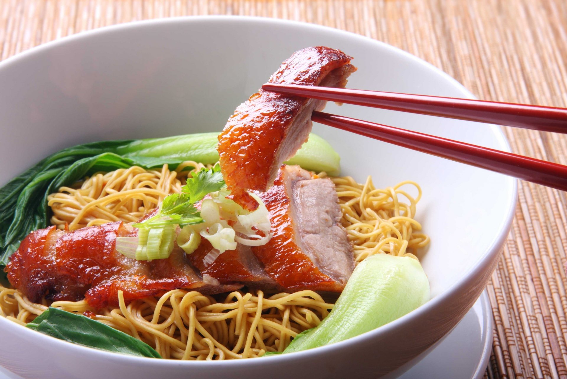 HD PC desktop wallpaper featuring a bowl of noodles with roasted meat, green vegetables, and chopsticks holding a piece of meat, highlighting a delicious meal.