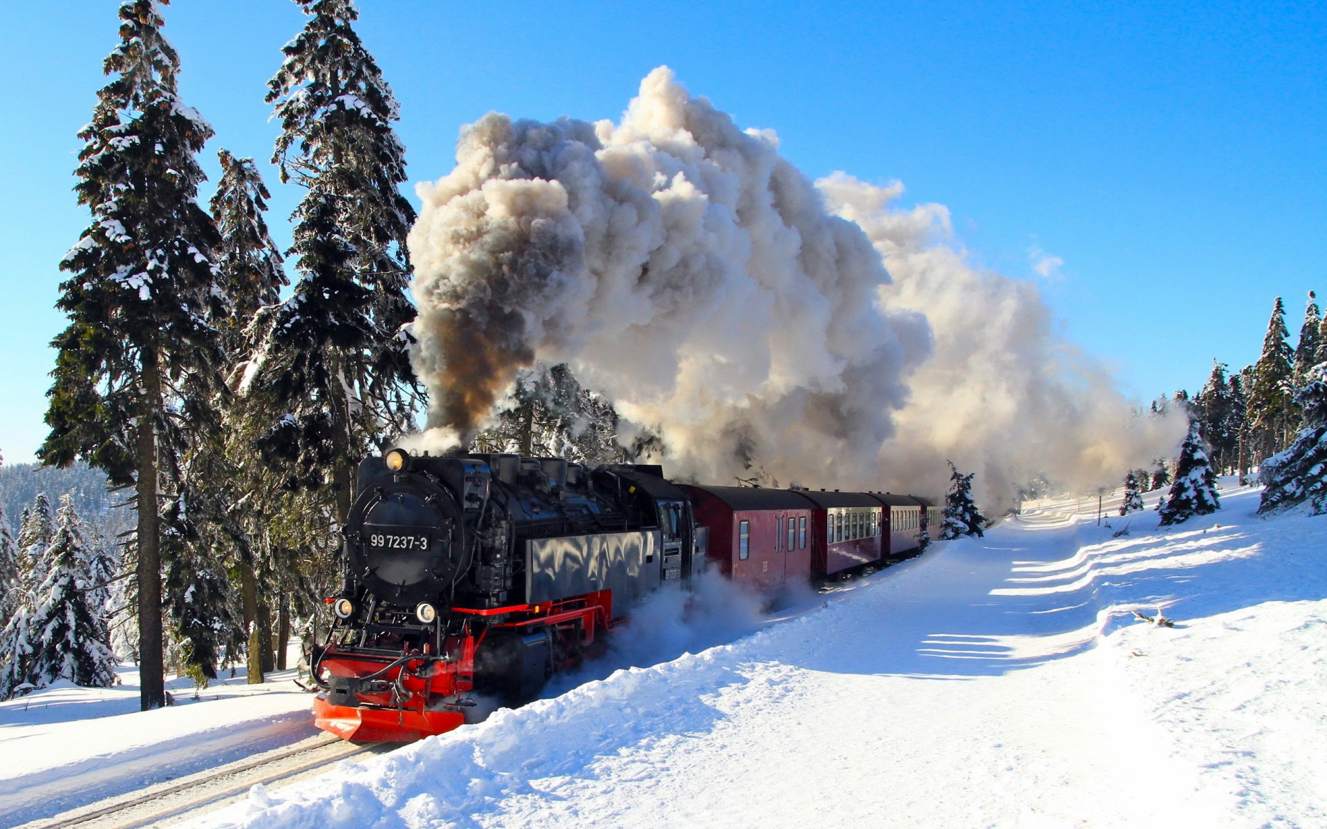 A vintage steam train billows smoke as it chugs through a snowy landscape, surrounded by tall evergreen trees, creating a stunning HD desktop wallpaper.
