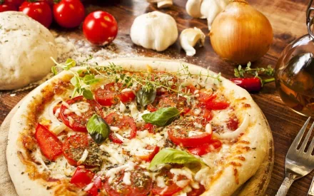 HD PC desktop wallpaper featuring a freshly baked pizza topped with tomatoes, basil, and herbs, surrounded by ingredients like garlic, onion, dough, and cherry tomatoes.