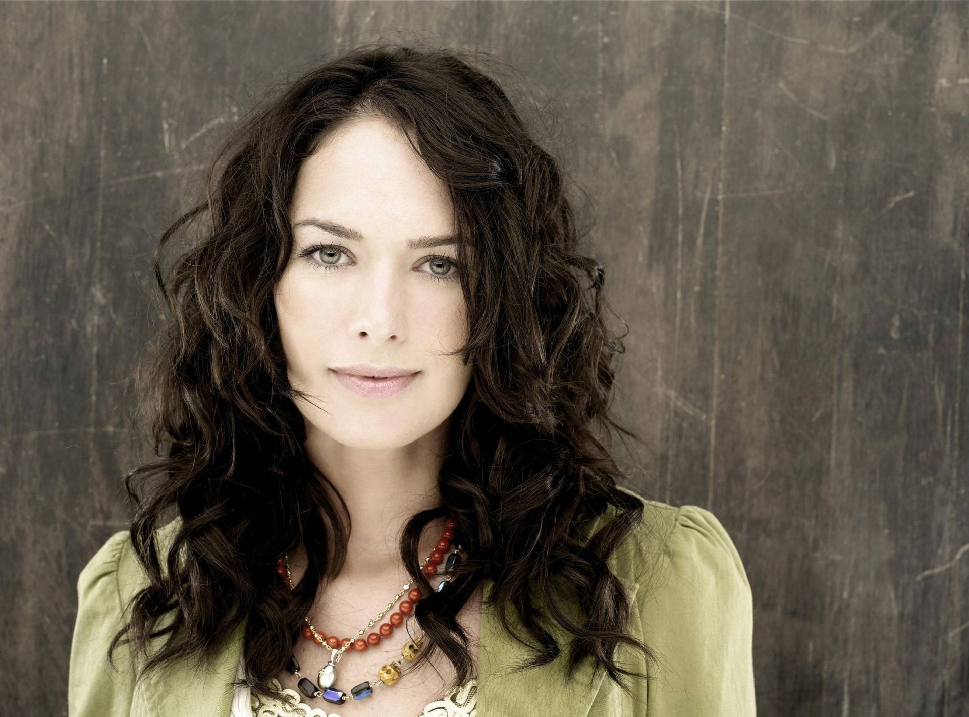 HD desktop wallpaper featuring celebrity Lena Headey with curly dark hair and a soft expression against a neutral textured background.