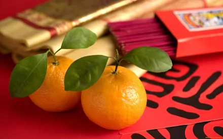 HD desktop wallpaper of two vibrant orange mandarins with green leaves resting on traditional red Chinese decorations, blending food and cultural elements.