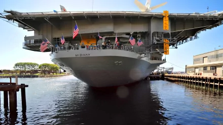 A striking HD wallpaper featuring a military aircraft carrier, the USS Midway, docked in serene waters with flags fluttering under a clear blue sky.