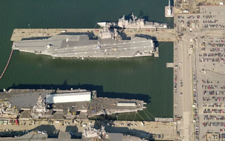 Aerial view of a military aircraft carrier docked alongside other vessels and a port, showcasing the intricate layout of naval operations. High-definition wallpaper for desktop use.