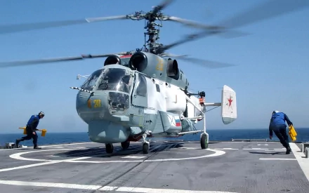 A Kamov Ka-27 military helicopter takes off from a ship's deck, with crew members preparing for flight against a clear blue sky, creating a dynamic HD desktop wallpaper.