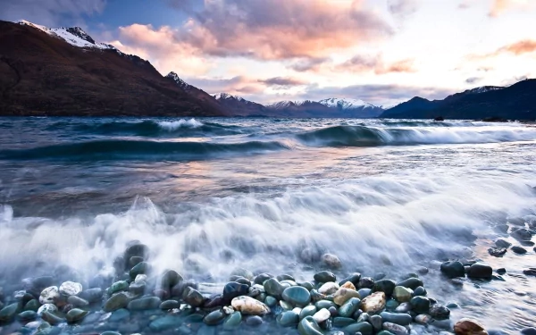 A scenic view of waves gently crashing over pebbles, with majestic mountains in the background and a colorful sky at dusk, capturing the beauty of nature and the ocean.
