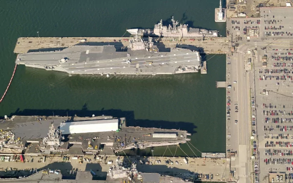 Aerial view of a military aircraft carrier docked alongside other vessels and a port, showcasing the intricate layout of naval operations. High-definition wallpaper for desktop use.