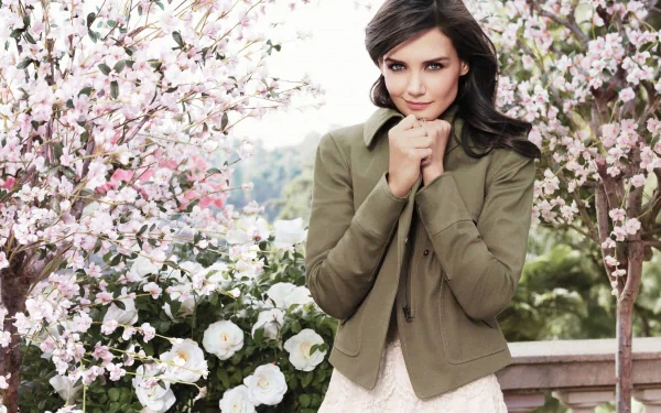 HD desktop wallpaper featuring American actress Katie Holmes with blue eyes and black hair, standing among blooming pink and white flowers.