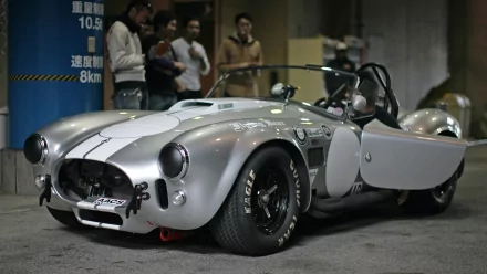 HD PC desktop wallpaper featuring a classic silver AC Cobra sports car with its door open, showcased in an indoor setting with people in the background.