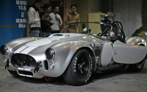 HD PC desktop wallpaper featuring a classic silver AC Cobra sports car with its door open, showcased in an indoor setting with people in the background.