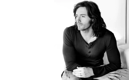 Black and white image of celebrity Hugh Jackman, gazing thoughtfully while seated, showcasing a confident and introspective expression. A stylish HD wallpaper for desktop use.