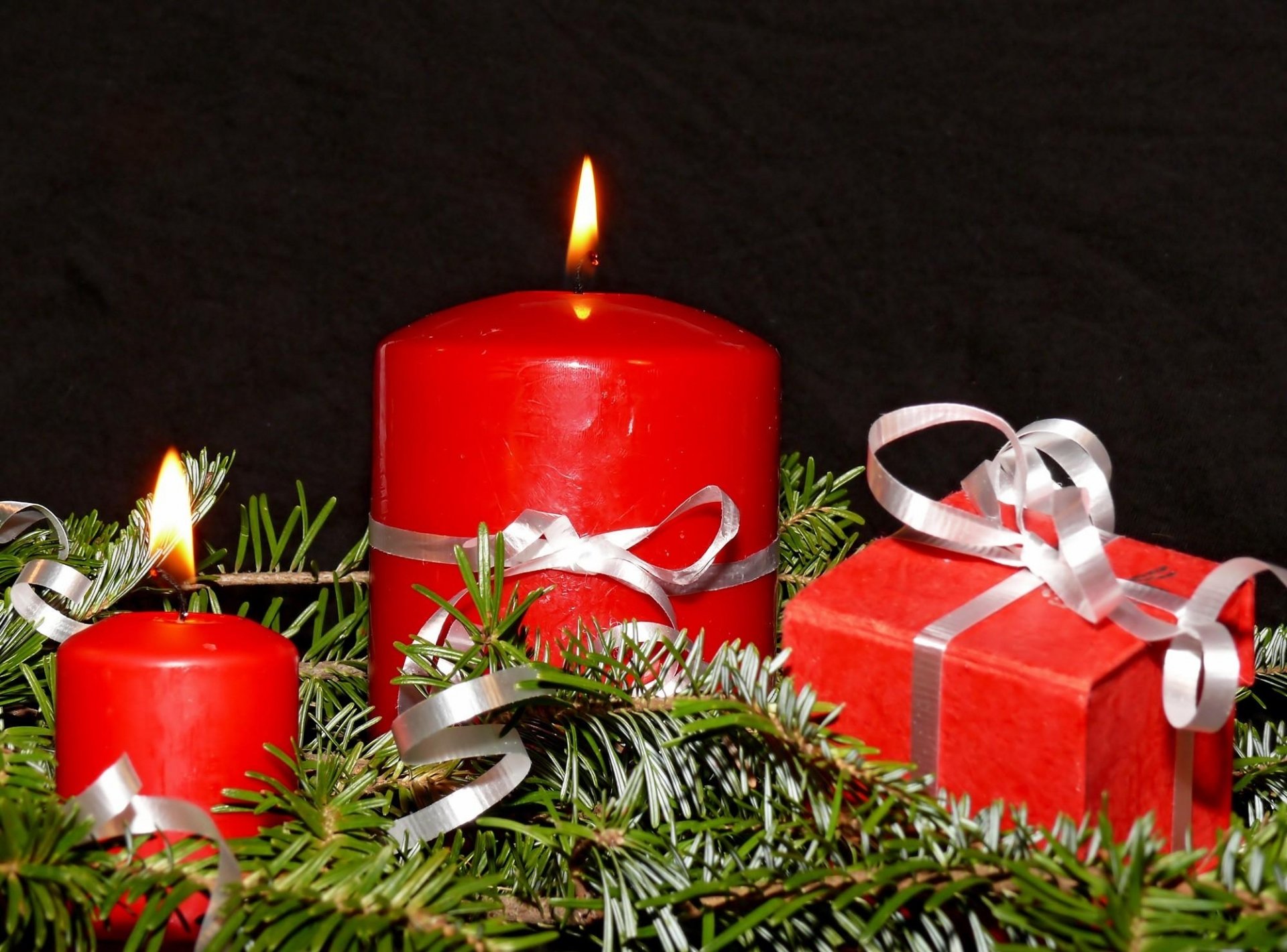 HD desktop wallpaper featuring two lit red candles, a red gift box with white ribbon, and festive green pine branches, evoking a warm Christmas holiday atmosphere.