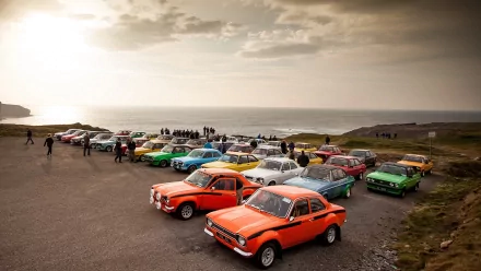 Vehicle lineup featuring classic Ford Escort cars in bright colors parked by the sea, composed as a 2K Quad HD PC desktop wallpaper background.