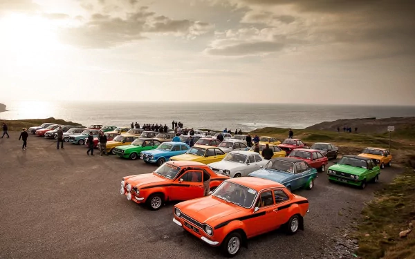 Vehicle lineup featuring classic Ford Escort cars in bright colors parked by the sea, composed as a 2K Quad HD PC desktop wallpaper background.