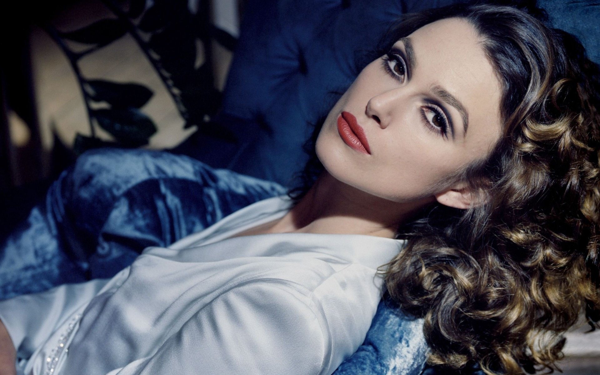 A captivating HD desktop wallpaper featuring celebrity Keira Knightley, showcasing her glamorous look with elegant curls and striking makeup against a plush blue background.