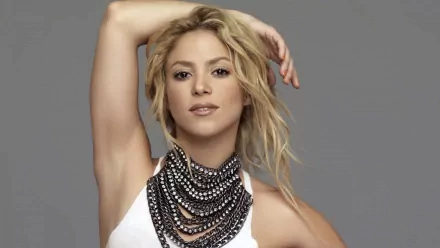 HD wallpaper of Shakira posing with one arm raised, wearing a stack of black and silver necklaces against a gray background. Keywords: music, Shakira.
