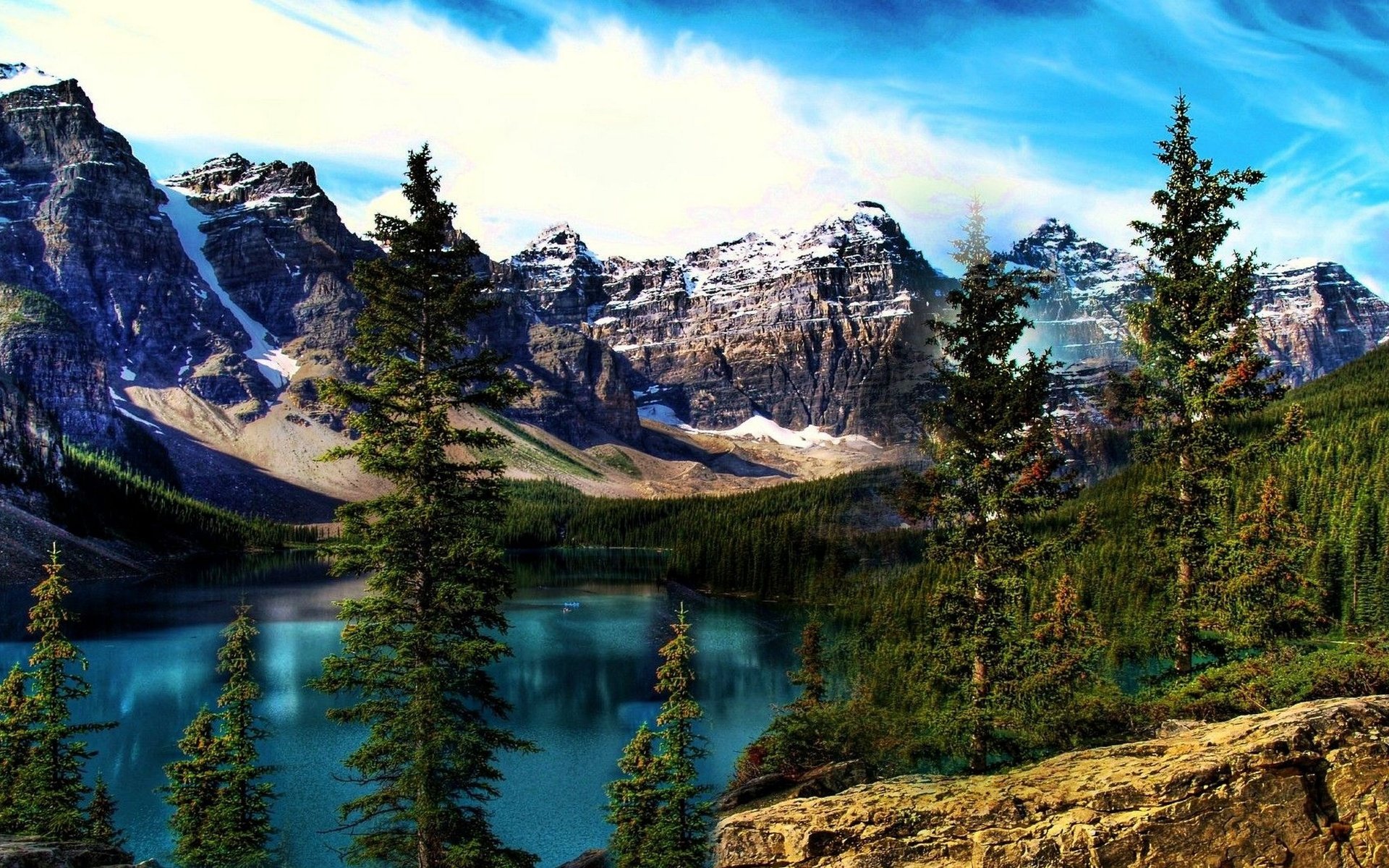 HD PC desktop wallpaper featuring a vibrant nature landscape with evergreen trees, a clear blue lake, and rugged snow-capped mountains under a bright sky.