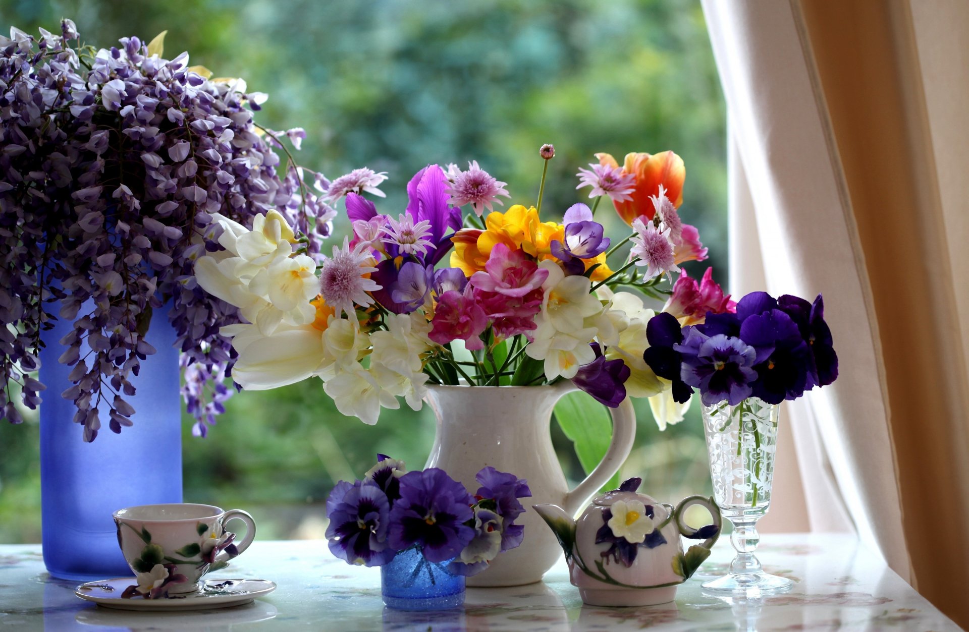 A vibrant still life arrangement featuring a variety of colorful flowers in a white vase, complemented by decorative teacups, against a soft-focus green background. HD photography.