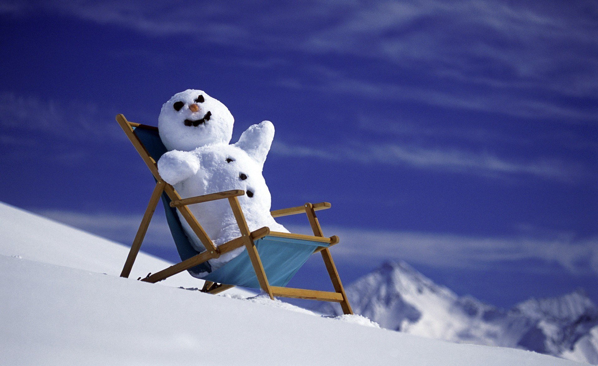 HD PC desktop wallpaper photography of a cheerful snowman reclining in a deck chair on a snowy slope with distant mountains and a deep blue sky.