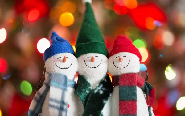 Three cheerful snowmen dressed in colorful hats and scarves stand together with a backdrop of bokeh Christmas lights. This HD image makes a festive desktop wallpaper and background.