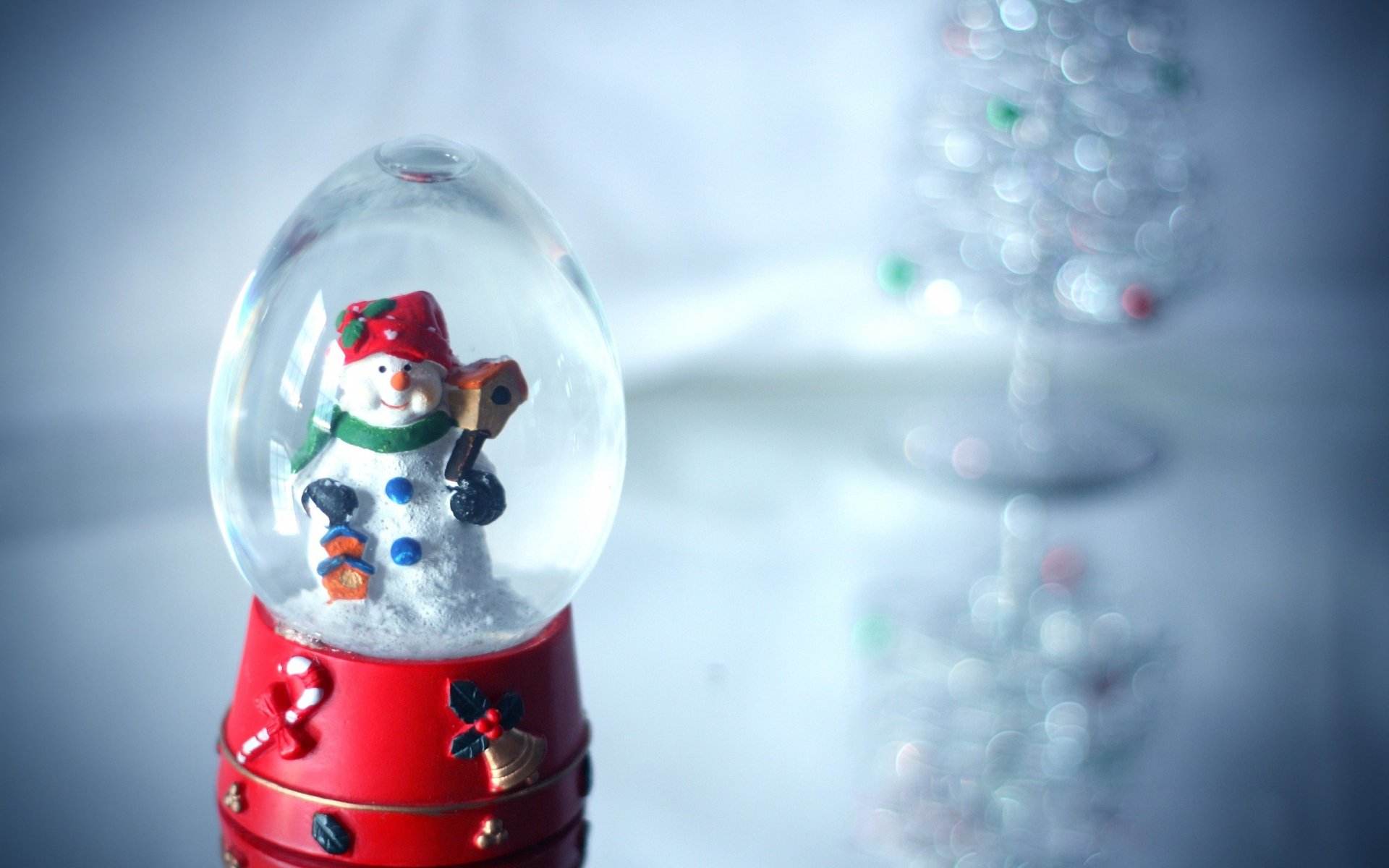 HD desktop wallpaper featuring a festive snow globe with a snowman inside, surrounded by softly falling snow and a blurred Christmas tree in the background.