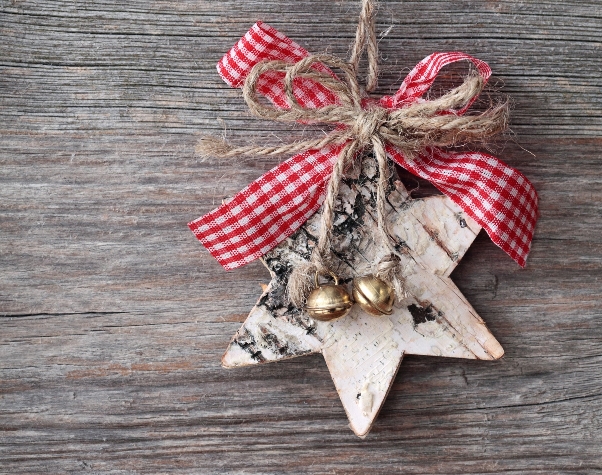 A rustic Christmas ornament shaped like a star, adorned with red and white checked ribbon and gold bells, set against a wooden background. A charming holiday desktop wallpaper.