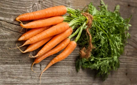 A fresh bunch of vibrant carrots with green tops rests on a rustic wooden surface, creating a colorful and appetizing HD desktop wallpaper.