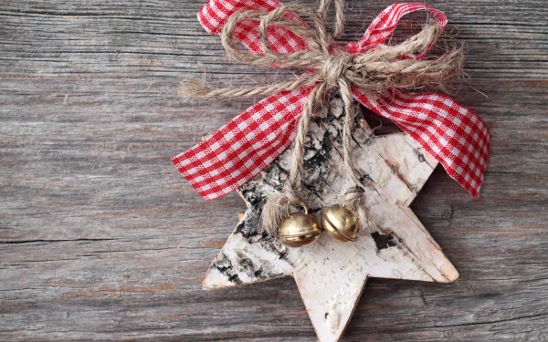 A rustic Christmas ornament shaped like a star, adorned with red and white checked ribbon and gold bells, set against a wooden background. A charming holiday desktop wallpaper.