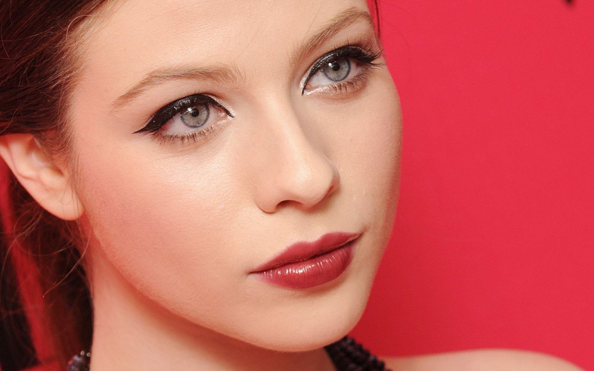 Close-up of celebrity Michelle Trachtenberg, featuring her striking blue eyes and bold makeup against a vibrant pink background, designed as a high-definition desktop wallpaper.