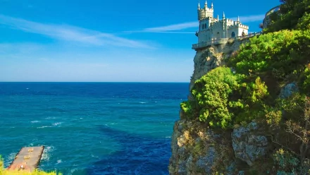 HD desktop wallpaper of Swallow's Nest, a castle perched on a cliff overlooking the sea in Ukraine, with the horizon in the background. The scene features lush greenery and a vivid, deep blue ocean.