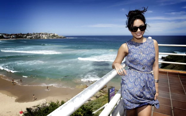 Vanessa Hudgens in a stylish blue dress and sunglasses, smiling by a seaside railing with ocean waves and a coastal town in the background, 4K Ultra HD wallpaper.
