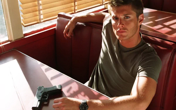 A high-definition wallpaper featuring Jensen Ackles seated at a booth, looking directly at the camera with a handgun resting on the table, illuminated by soft sunlight.