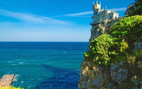 HD desktop wallpaper of Swallow's Nest, a castle perched on a cliff overlooking the sea in Ukraine, with the horizon in the background. The scene features lush greenery and a vivid, deep blue ocean.