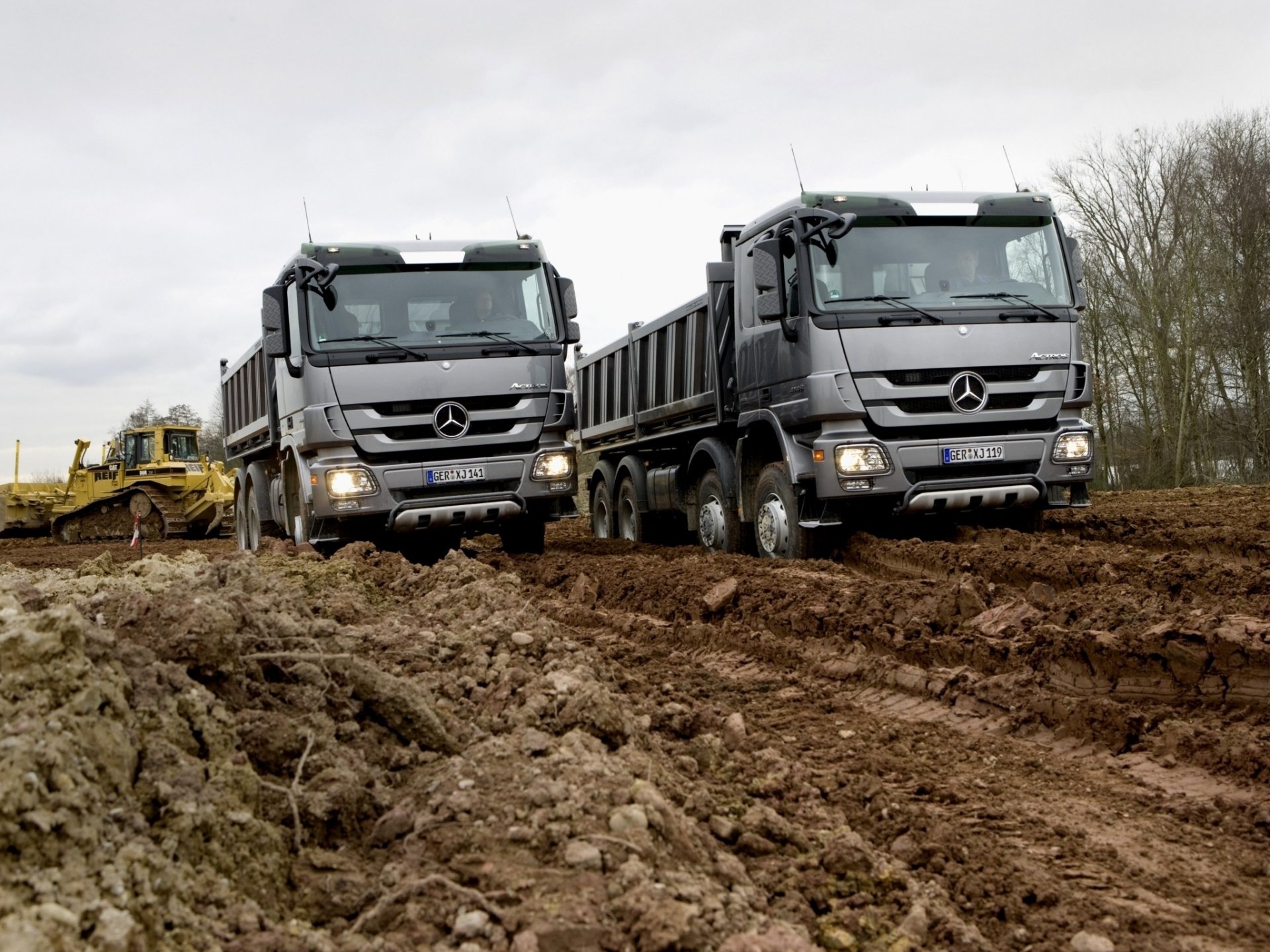 HD PC desktop wallpaper featuring two Mercedes dump trucks navigating a muddy construction site under an overcast sky.