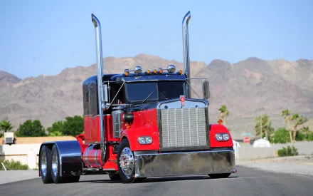 A striking Kenworth truck in vibrant red, showcasing its impressive design with tall exhaust stacks and chrome accents, drives along a desert road, capturing a bold, adventurous spirit.