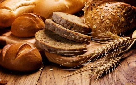 A close-up of freshly baked bread, including sliced whole grain and crusty rolls, surrounded by wheat stalks on a rustic wooden surface, serving as a vibrant desktop wallpaper.