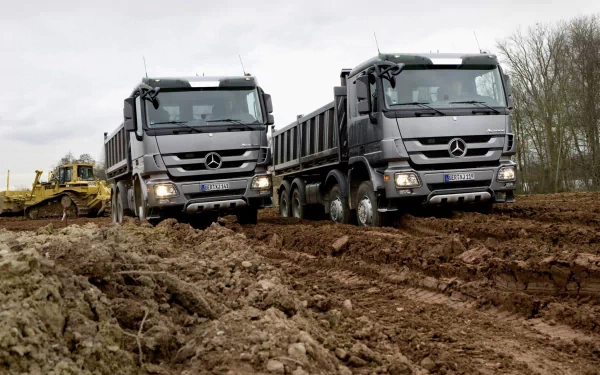HD PC desktop wallpaper featuring two Mercedes dump trucks navigating a muddy construction site under an overcast sky.