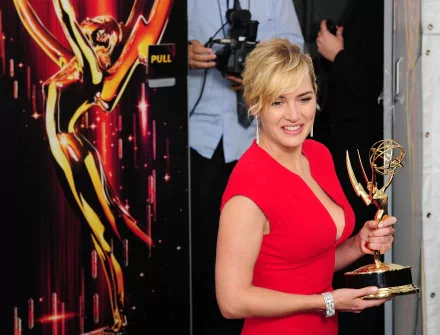 Kate Winslet smiles while holding an Emmy award, dressed in a vibrant red dress, with a backdrop featuring a glamorous Hollywood theme. A striking HD desktop wallpaper.