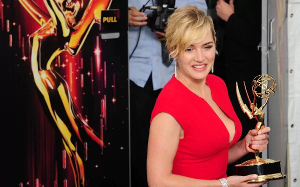 Kate Winslet smiles while holding an Emmy award, dressed in a vibrant red dress, with a backdrop featuring a glamorous Hollywood theme. A striking HD desktop wallpaper.