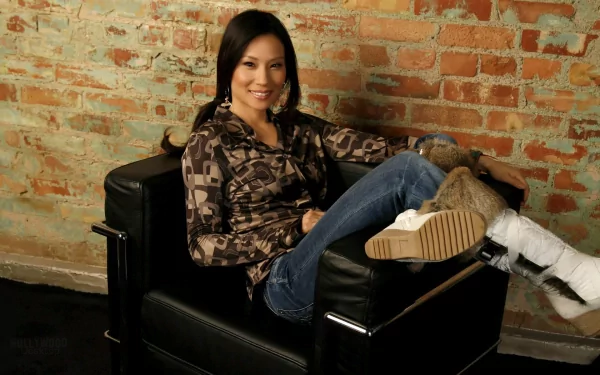 Lucy Liu lounges comfortably in a stylish black chair, showcasing a casual look against a textured brick background. This HD desktop wallpaper captures her relaxed vibe.