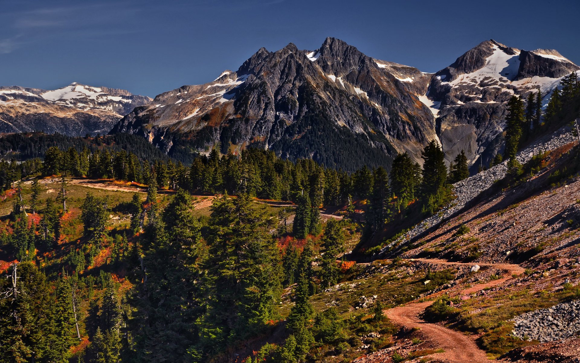 HD PC desktop wallpaper showcasing a rugged mountain range with snow-capped peaks, dense evergreen forests, and a winding dirt path under a clear blue sky.