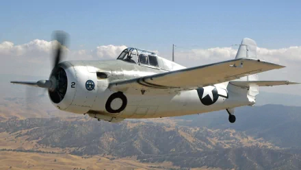 HD desktop wallpaper featuring a Grumman F4F Wildcat military aircraft flying over a mountainous landscape under a blue sky.