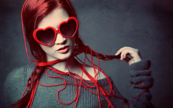 A woman with vibrant red hair styled in braids wears oversized heart-shaped sunglasses. She poses confidently against a muted background, creating a striking HD desktop wallpaper.