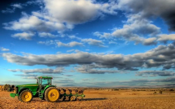 A John Deere 9120 tractor stands prominently in a vast farm landscape, under a sky filled with fluffy clouds, showcasing the beauty of rural agriculture.