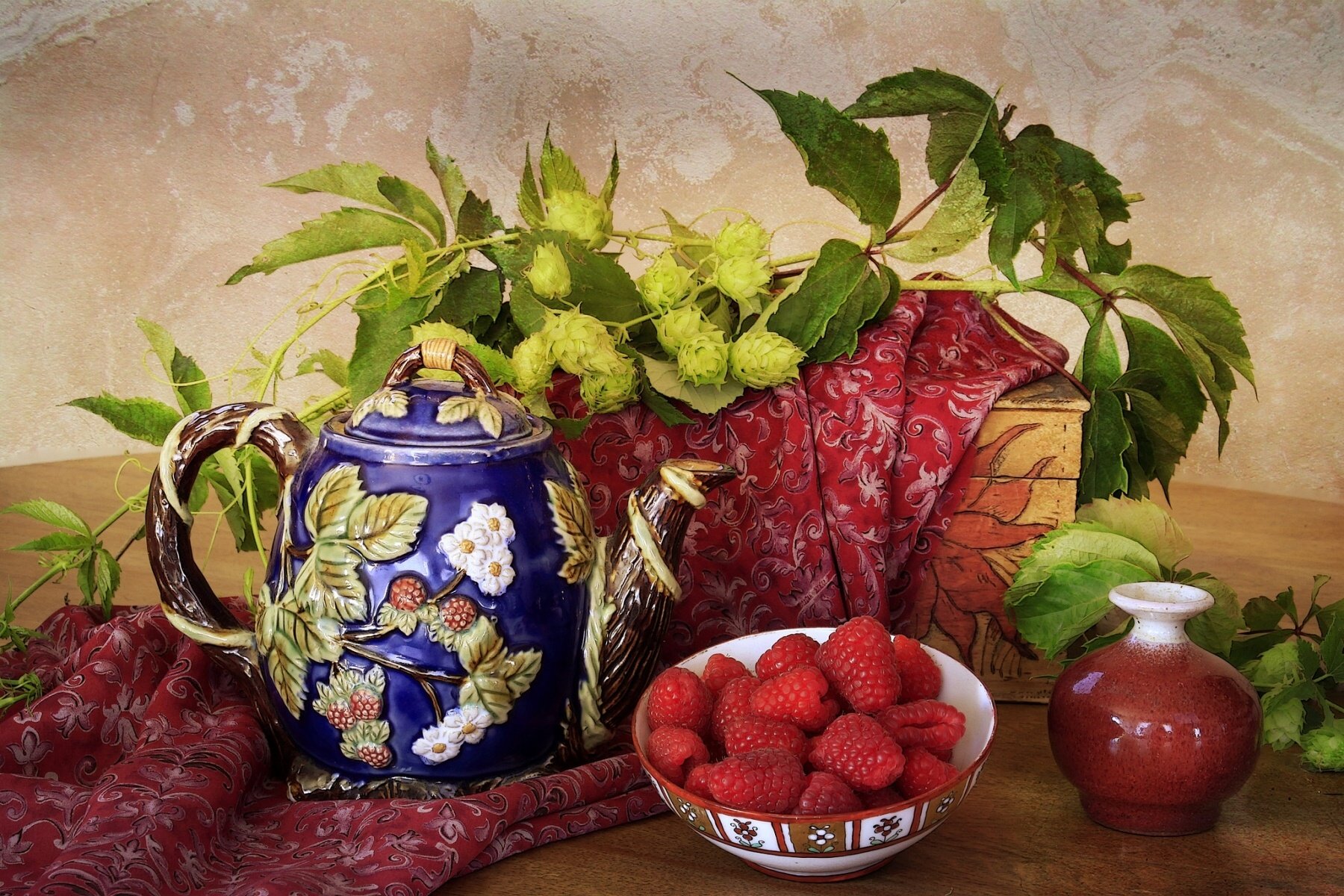HD desktop wallpaper still life: ornate blue teapot, bowl of ripe raspberries, small red vase and leafy branches arranged on a patterned cloth.