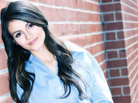 A high-definition desktop wallpaper featuring Victoria Justice, smiling softly against a textured brick wall, showcasing her long, flowing hair and casual style.